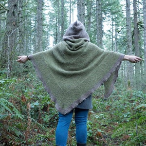 May include: A hooded green and brown knitted poncho with a fringed edge. The poncho is worn by a person standing in a forest.