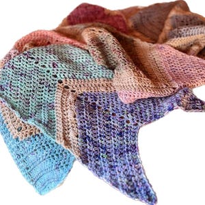 May include: A triangular crochet shawl featuring a variegated color palette of light blue, mint green, soft pink, and deep purple. The textile displays a textured stitch pattern with subtle speckles throughout the yarn, creating a soft gradient.