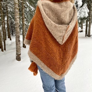May include: A hooded knitted poncho with a brown and beige color scheme. The poncho is made of a soft, fuzzy yarn and has a triangular shape. The person wearing the poncho is standing in a snowy forest.