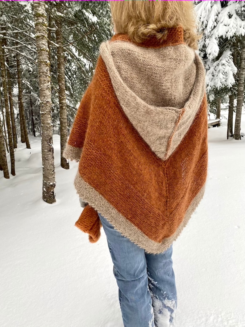 May include: A hooded knitted poncho with a brown and beige color scheme. The poncho is made of a soft, fuzzy yarn and has a triangular shape. The person wearing the poncho is standing in a snowy forest.
