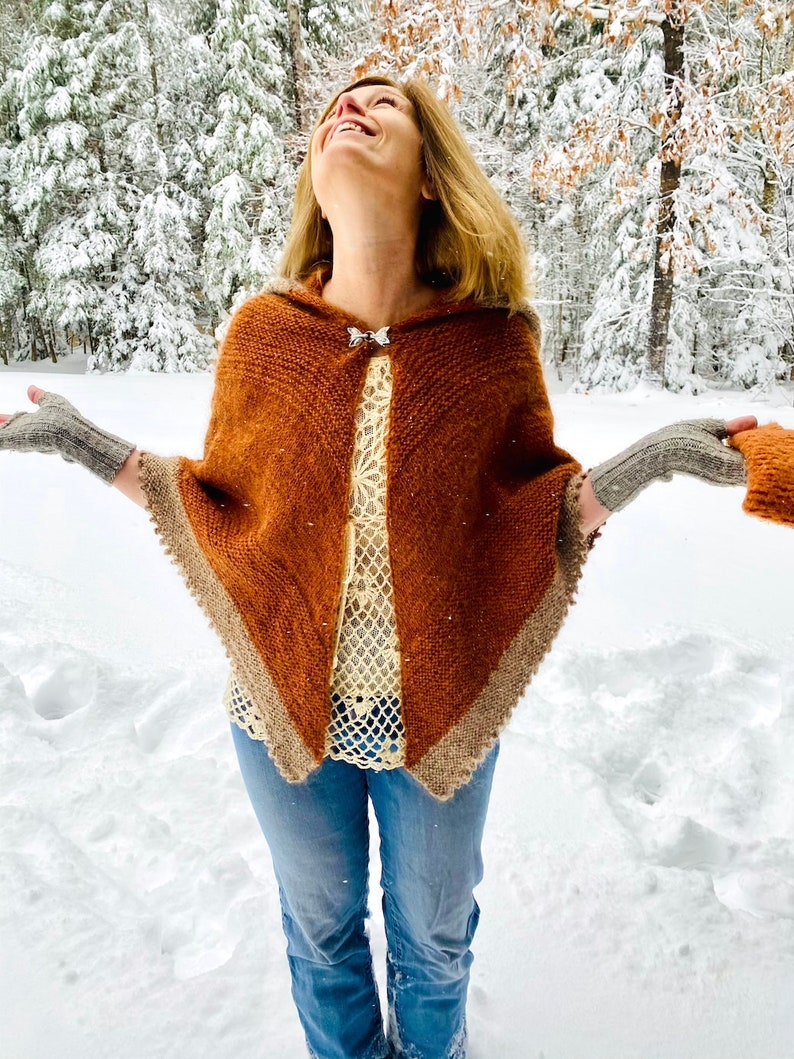 May include: A woman wearing a brown hooded poncho with a white crocheted top and blue jeans. She is standing in a snowy forest and smiling.