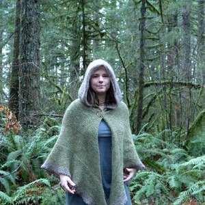 May include: A woman wearing a green hooded knitted cape with a brown trim in a forest setting.
