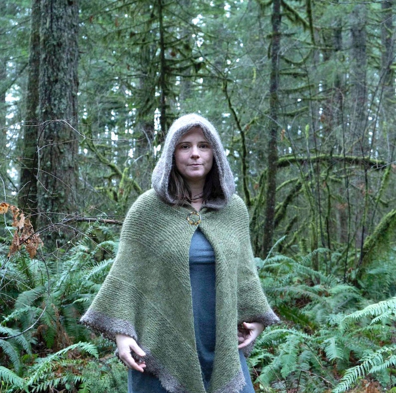 May include: A woman wearing a green hooded knitted cape with a brown trim in a forest setting.
