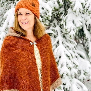 May include: A woman wearing a brown knitted hooded shawl with a silver pin, a brown knit hat, and a white lace top. She is standing in front of a snowy evergreen tree.