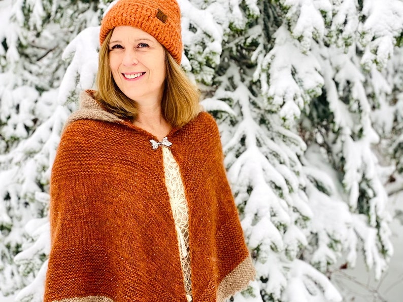 May include: A woman wearing a brown knitted hooded shawl with a silver pin, a brown knit hat, and a white lace top. She is standing in front of a snowy evergreen tree.