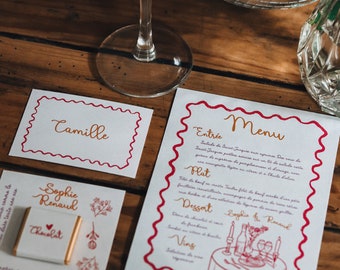 Puede incluir: Un menú de boda con un borde rojo y letras doradas. El menú incluye una ensalada, un plato de carne de res, postre y vino. El menú está sobre una mesa de madera con un vaso de jugo de naranja y una barra de chocolate con un corazón.