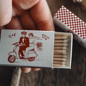 Peut inclure: Une boîte d'allumettes avec un motif à carreaux rouges et blancs sur le manchon extérieur. La boîte d'allumettes présente une illustration rouge d'un couple sur un scooter avec le texte "C+F" et "Dolce Vita". Les allumettes sont visibles à l'intérieur de la boîte.