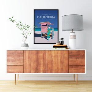 May include: A framed poster with a blue background and the text "Welcome to California". The poster features a pink lifeguard stand with a surfboard in front of it. The poster is hanging on a white wall above a wooden dresser with a lamp and a plant.