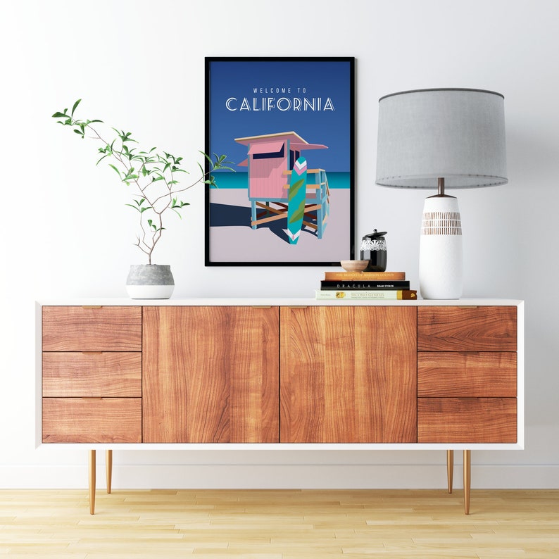May include: A framed poster with a blue background and the text "Welcome to California". The poster features a pink lifeguard stand with a surfboard in front of it. The poster is hanging on a white wall above a wooden dresser with a lamp and a plant.