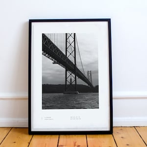 May include: Black and white photograph of the 25 de Abril Bridge in Lisbon, Portugal. The bridge is a suspension bridge that crosses the Tagus River. The photo is taken from below the bridge, looking up at the structure.