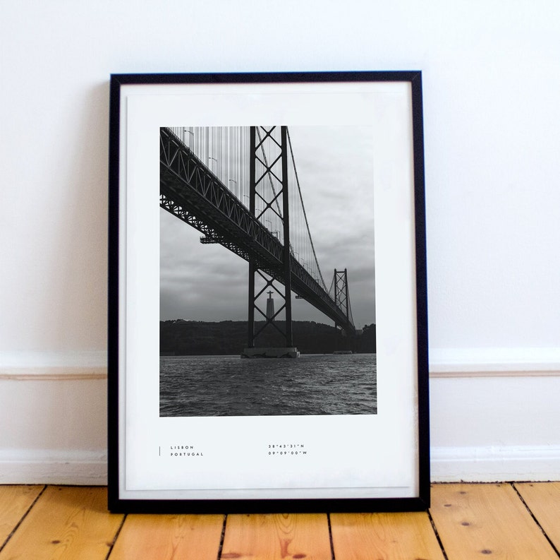 May include: Black and white photograph of the 25 de Abril Bridge in Lisbon, Portugal. The bridge is a suspension bridge that crosses the Tagus River. The photo is taken from below the bridge, looking up at the structure.
