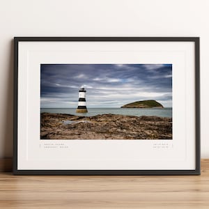 May include: Framed print of Puffin Island, Anglesey, Wales. The image features a black and white striped lighthouse on a rocky shore, with a small island in the background under a cloudy sky. The print is in a black frame and rests on a wooden surface.