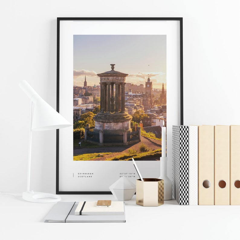 May include: A framed photograph of a cityscape with a monument in the foreground. The city is Edinburgh, Scotland. The monument is the Scott Monument, a neo-Gothic structure built in honor of Sir Walter Scott. The photograph is taken from Calton Hill, a hill overlooking the city. The sky is a soft orange and pink, indicating sunset. The text on the photograph reads "EDINBURGH SCOTLAND 55&deg;57'10"N 03&deg;11'20"W".