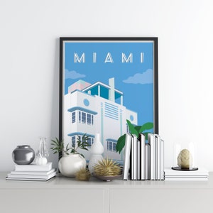 May include: A framed print of a Miami Art Deco building with the word "MIAMI" in white letters on a blue background. The print is displayed on a white shelf with a variety of decorative objects, including vases, books, and a glass dome with a decorative object inside.