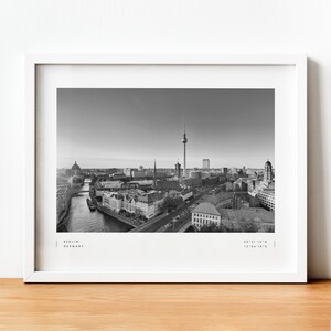 Puede incluir: Impresi&oacute;n en blanco y negro enmarcada de Berl&iacute;n, Alemania, con el horizonte de la ciudad y la Torre de Televisi&oacute;n como punto focal. La impresi&oacute;n incluye el nombre de la ciudad y las coordenadas. El marco es blanco, sobre una superficie de madera.