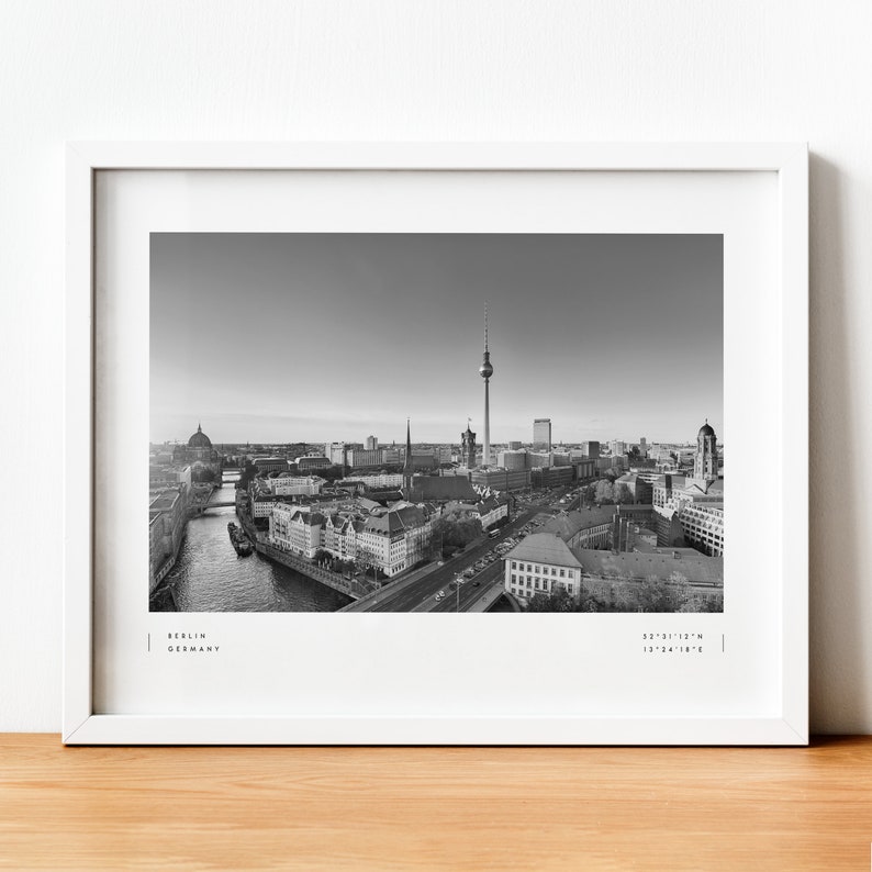 Puede incluir: Impresi&oacute;n en blanco y negro enmarcada de Berl&iacute;n, Alemania, con el horizonte de la ciudad y la Torre de Televisi&oacute;n como punto focal. La impresi&oacute;n incluye el nombre de la ciudad y las coordenadas. El marco es blanco, sobre una superficie de madera.