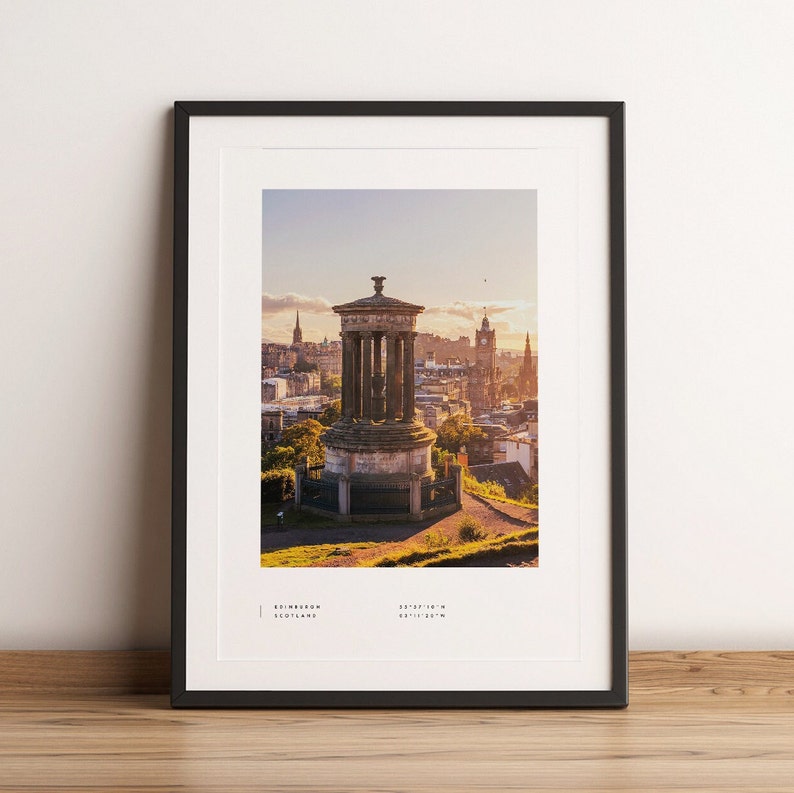 May include: A framed photograph of a stone monument on a hill overlooking the city of Edinburgh, Scotland. The monument is a tall, cylindrical structure with a domed roof. The city is in the background, with buildings and trees visible. The sky is a light blue with some clouds. The photo is taken from a high angle, looking down on the city.