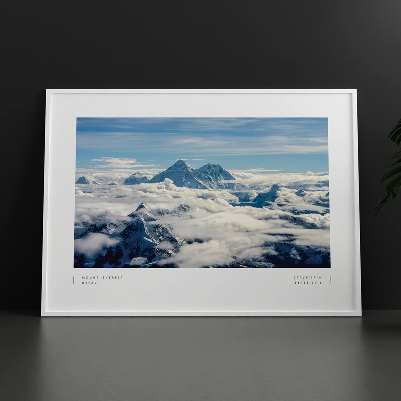 Pu&ograve; includere: Una vista panoramica del Monte Everest, la montagna pi&ugrave; alta del mondo, con cime innevate che si ergono sopra un mare di nuvole. Il cielo &egrave; di un blu brillante con nuvole bianche e vaporose. Il testo "MOUNT EVEREST, NEPAL" e le coordinate "27&deg;59'17"N 86&deg;55'31"E" sono stampate nella parte inferiore dell'immagine.