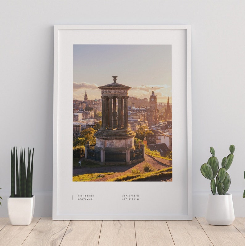 May include: A framed photograph of a stone monument overlooking a city skyline. The monument is a tall, cylindrical structure with a domed roof. The city skyline is in the background, with buildings and trees visible. The photograph is taken from a high vantage point, and the sun is setting in the distance, casting a warm glow over the scene. The text "EDINBURGH, SCOTLAND 55.9521&deg;N 3.1881&deg;W" is printed at the bottom of the photograph.