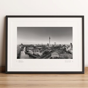 Puede incluir: Impresi&oacute;n enmarcada en blanco y negro de Berl&iacute;n, Alemania, con el paisaje urbano, la Torre de Televisi&oacute;n y el r&iacute;o Spree. La impresi&oacute;n incluye el nombre de la ciudad y las coordenadas. El marco es negro y se muestra sobre madera.