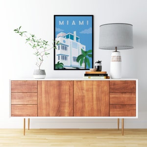 May include: A framed illustration of a white building with a blue sky and palm trees. The illustration is titled "MIAMI". The framed illustration is on a white shelf with a lamp and books.