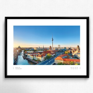 Puede incluir: Fotograf&iacute;a panor&aacute;mica enmarcada de Berl&iacute;n, Alemania, con la ic&oacute;nica Torre de Televisi&oacute;n, el r&iacute;o Spree y arquitectura hist&oacute;rica bajo un cielo azul claro. La impresi&oacute;n incluye el nombre de la ciudad y sus coordenadas.