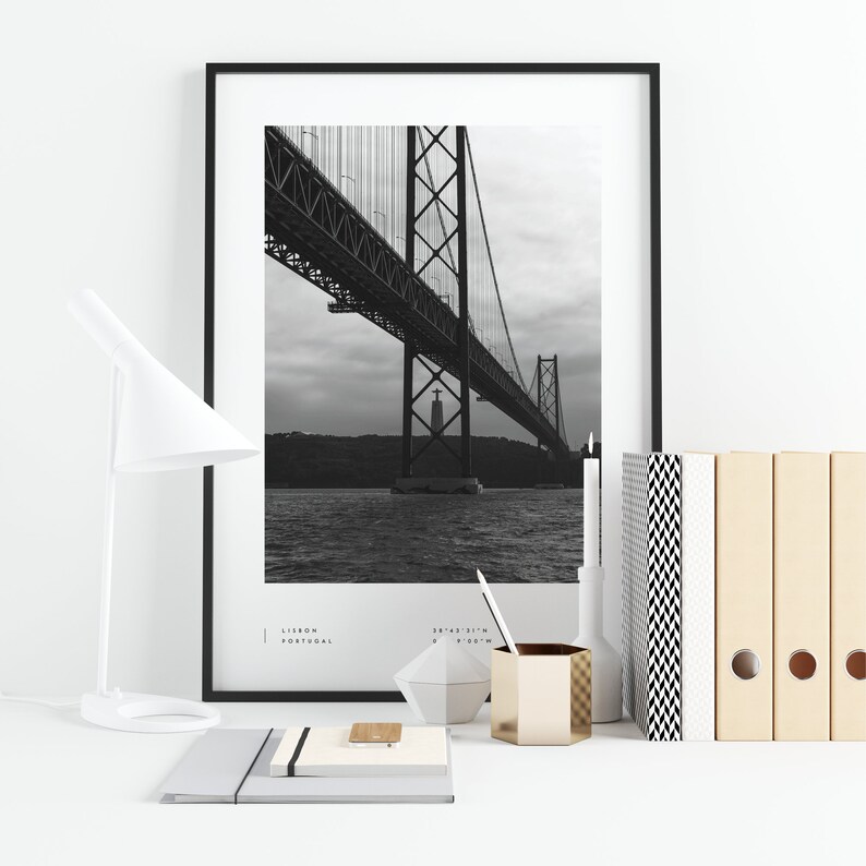 May include: Black and white photograph of a bridge in Lisbon, Portugal. The bridge is a suspension bridge with a large tower in the center. The photo is framed in a black frame.