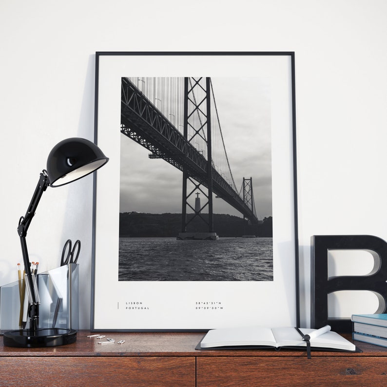 May include: Black and white photograph of the 25 de Abril Bridge in Lisbon, Portugal. The bridge is a suspension bridge with a large tower in the center. The photo is framed in a black frame.
