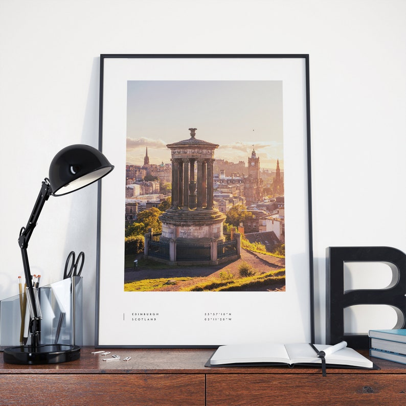 May include: A black and white photograph of a stone monument in Edinburgh, Scotland. The monument is on a hill overlooking the city. The city is in the background and the sky is a light blue color. The text "EDINBURGH SCOTLAND" and coordinates are at the bottom of the image.