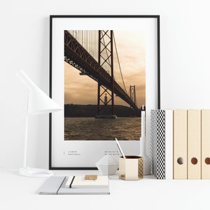 May include: A framed photograph of the 25 de Abril Bridge in Lisbon, Portugal. The bridge is a suspension bridge with a red steel structure and is seen from below. The photo is taken at sunset, with the sky a warm orange and yellow. The text "LISBON, PORTUGAL" and coordinates are printed below the image.