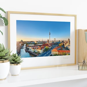 Puede incluir: Impresi&oacute;n enmarcada de Berl&iacute;n, Alemania, con la Torre de la Televisi&oacute;n y el r&iacute;o Spree. El marco dorado se exhibe en una estanter&iacute;a blanca con plantas y un revistero de madera. Coordenadas en la parte inferior.