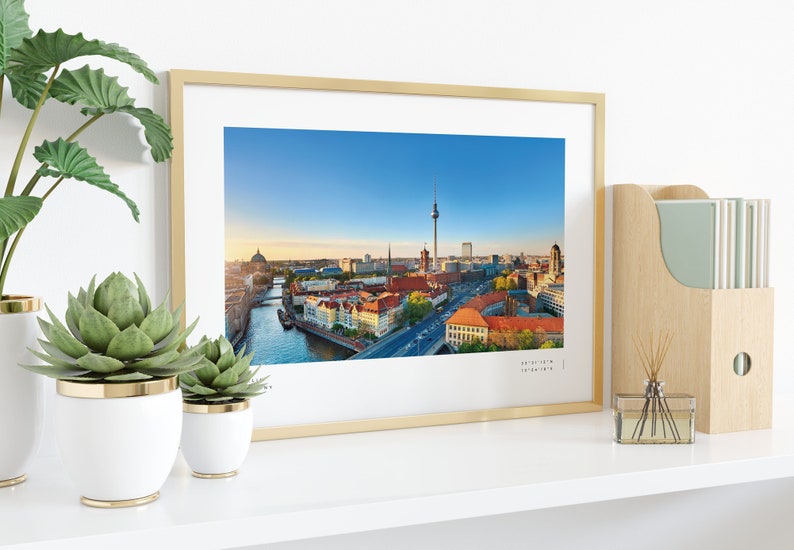 Puede incluir: Impresi&oacute;n enmarcada de Berl&iacute;n, Alemania, con la Torre de la Televisi&oacute;n y el r&iacute;o Spree. El marco dorado se exhibe en una estanter&iacute;a blanca con plantas y un revistero de madera. Coordenadas en la parte inferior.