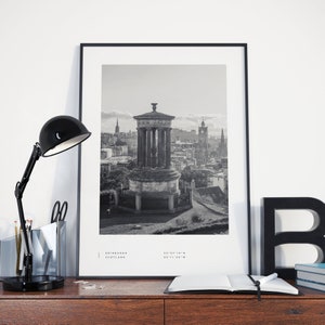 May include: Black and white photograph of the Edinburgh skyline featuring a monument with a columned structure and a domed roof. The photo is framed in a black frame and is displayed on a wooden surface with a black lamp, a notebook, and a black letter 'B' on the right side of the image. The text 'EDINBURGH SCOTLAND 55&deg;57'10"N 03&deg;11'20"W' is printed at the bottom of the photo.