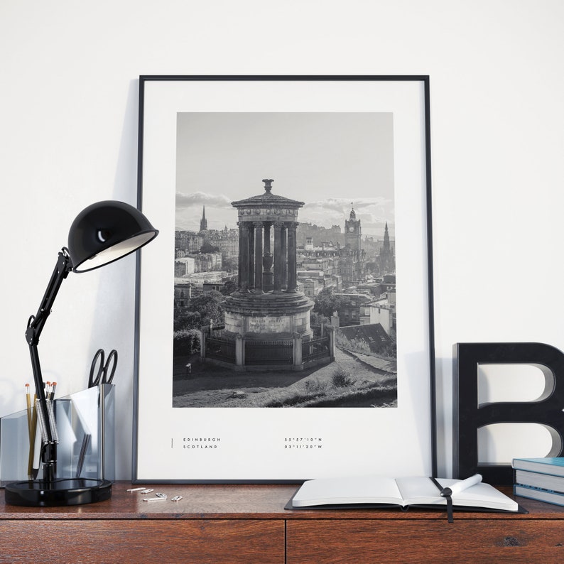 May include: Black and white photograph of the Edinburgh skyline featuring a monument with a columned structure and a domed roof. The photo is framed in a black frame and is displayed on a wooden surface with a black lamp, a notebook, and a black letter 'B' on the right side of the image. The text 'EDINBURGH SCOTLAND 55&deg;57'10"N 03&deg;11'20"W' is printed at the bottom of the photo.