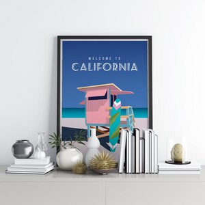 May include: A framed print with a blue background and a pink lifeguard stand with a surfboard leaning against it. The text "WELCOME TO CALIFORNIA" is at the top of the print.