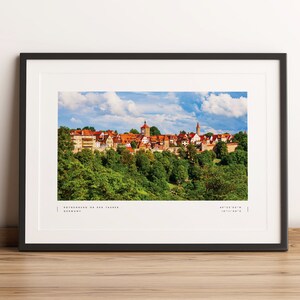 May include: A panoramic view of the town of Rothenburg ob der Tauber, Germany. The town is nestled in a valley and surrounded by lush green trees. The buildings are a mix of red tile roofs and white walls, with a few towers and spires visible.