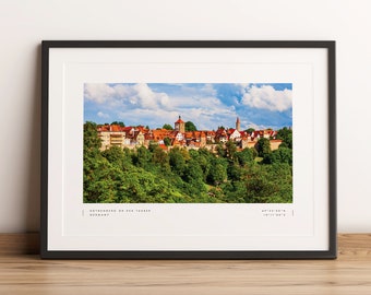 Puede incluir: Una vista panorámica de la ciudad de Rothenburg ob der Tauber, Alemania. La ciudad está enclavada en un valle y rodeada de exuberantes árboles verdes. Los edificios son una mezcla de tejados de tejas rojas y paredes blancas, con algunas torres y chapiteles visibles.