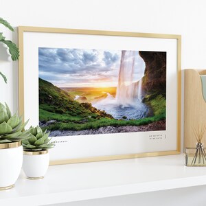 Op de afbeelding: Een ingelijste foto van een waterval die over een klif stroomt met een zonsondergang op de achtergrond. De foto is in een gouden lijst en staat op een witte plank met planten in witte potten.