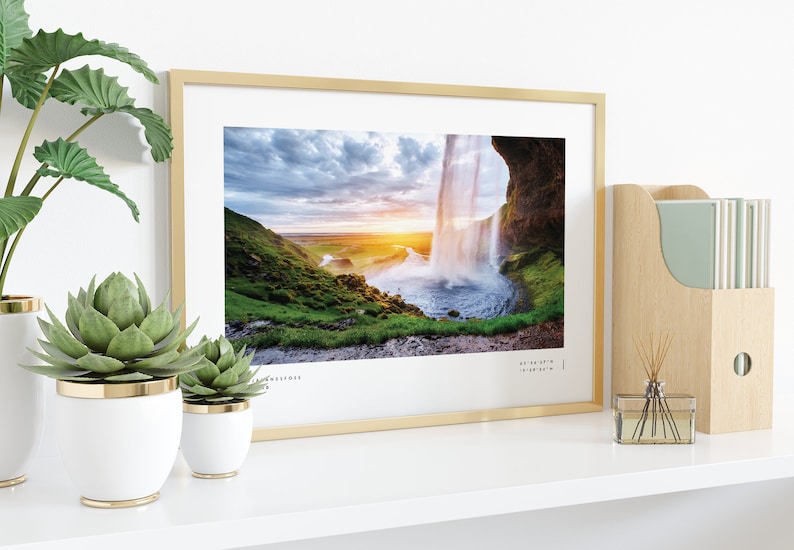 Op de afbeelding: Een ingelijste foto van een waterval die over een klif stroomt met een zonsondergang op de achtergrond. De foto is in een gouden lijst en staat op een witte plank met planten in witte potten.