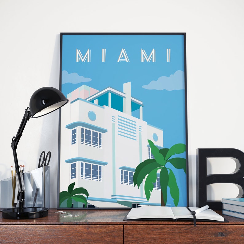 May include: A framed poster with a blue background and the word "MIAMI" in white letters. The poster features a white art deco building with a blue roof and palm trees in front of it.