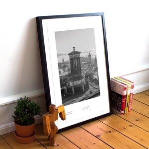 May include: Black and white photograph of a cityscape with a tall, round building in the center. The building has a dome and a columned structure. The city is in the background and is made up of many buildings. The photo is framed in a black frame.