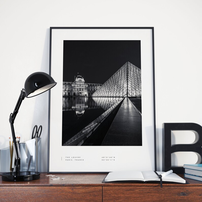 May include: Black and white photograph of the Louvre Museum in Paris, France at night. The iconic glass pyramid is reflected in the water in front of the building. The text "THE LOUVRE PARIS, FRANCE" is at the bottom of the image.