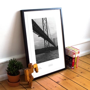 May include: Black and white photograph of a bridge over water. The bridge is a suspension bridge with a large tower in the center. The photo is framed in a black frame.