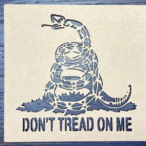 May include: A laser-cut wooden stencil featuring a coiled rattlesnake with its head raised. The words "DON'T TREAD ON ME" are cut out below the snake. The stencil is light beige, and the background is a dark blue.
