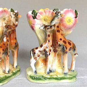 May include: Two ceramic vases, each featuring a giraffe family. The vases have a light yellow base with a pink and white flower design at the top. The giraffes are brown with dark brown spots.