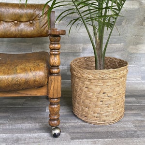 Puede incluir: Un sillón vintage de cuero marrón con estructura de madera y detalles decorativos. Una cesta de mimbre tejida contiene una planta verde. La silla tiene respaldo y reposabrazos con mechones. La pata de la silla tiene una rueda plateada.