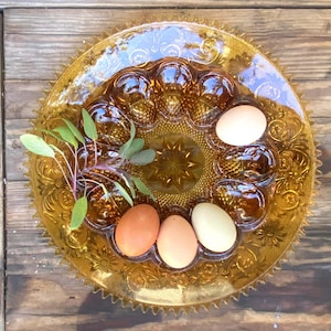 Peut inclure: Plat à œufs farcis en verre ambré avec un bord décoratif, contenant cinq œufs de couleurs variées. Des herbes vertes fraîches sont placées au centre. Le plat repose sur une surface en bois rustique.