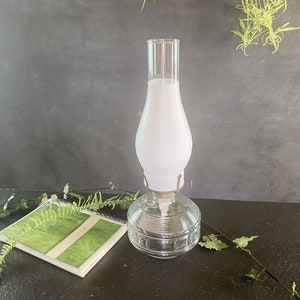 May include: A clear glass oil lamp featuring a round, ribbed base and a tall, frosted white glass chimney. The lamp includes a metal burner assembly with a small adjustment knob, designed to hold the glass shade securely in place.