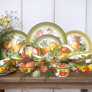 Pode incluir: Uma coleção de pratos de esmalte pintados à mão com um design de maçã vibrante. O conjunto inclui pratos, tigelas e recipientes com tampa em tons de verde, amarelo e branco. Um vaso de vidro verde com flores à esquerda.