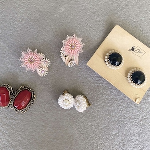 Könnte beinhalten: Eine Sammlung verschiedener Vintage-Ohrringe. Dazu gehören rosa Blüten-Clip-Ohrringe, rote rechteckige Steinohrringe, weiße perlenartige Clip-Ohrringe und schwarze Steinohrringe mit Perlenakzenten auf einer Vintage-Karte. Das Coco-Logo ist sichtbar.